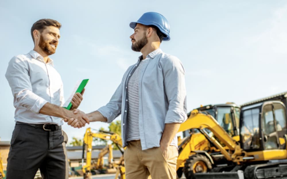 Two men shaking hands at a construction site with construction vehicles in the background.