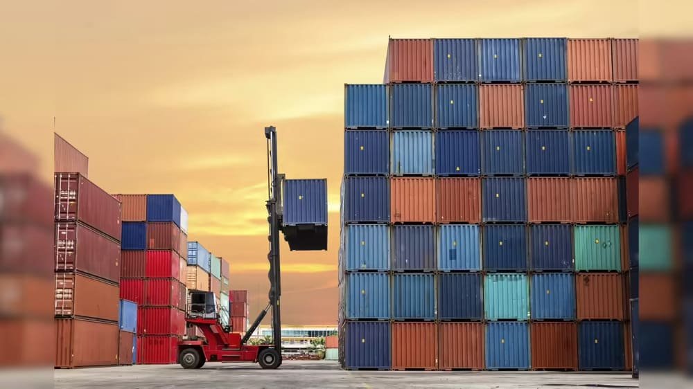 A forklift lifting a shipping container at a port during sunset, surrounded by stacks of red and blue containers