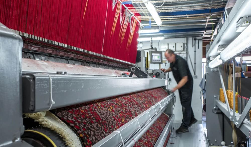 Factory setting with a large carpet weaving machine and a worker at a control panel
