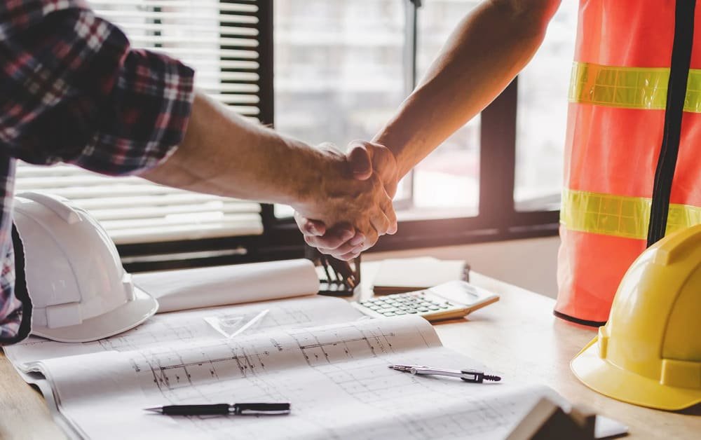 Two people shaking hands over a table with blueprints, helmets, and a calculator