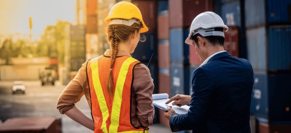 Two people wearing safety gear examine a document near stacked shipping containers