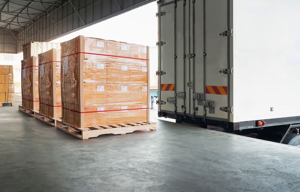 Pallets of cardboard boxes in a warehouse with a delivery truck at a loading dock