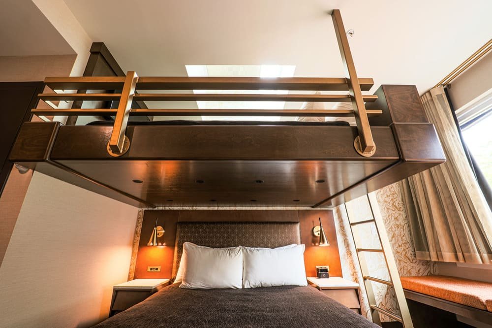 Loft-style bedroom with a wooden loft bed above a queen-sized bed, featuring a ladder and brass-colored lamps